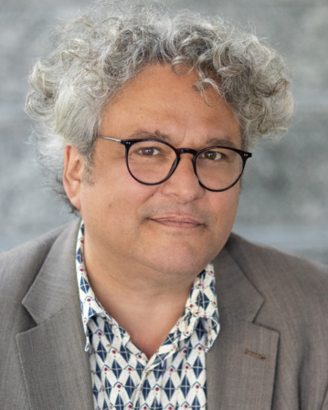 Ein Mann mit lockigem, grauem Haar und Brille trägt einen blauen Blazer über einem gemusterten Hemd. Er schaut direkt in die Kamera.