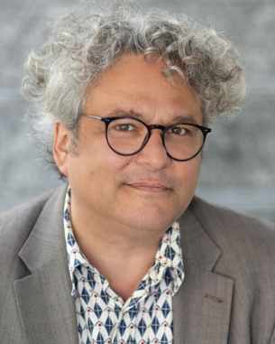 Ein Mann mit lockigem, grauem Haar und Brille trägt einen blauen Blazer über einem gemusterten Hemd. Er schaut direkt in die Kamera.