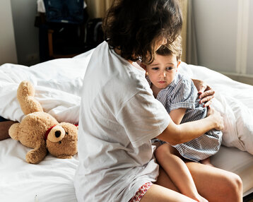 A person is hugging a child on a bed, while a teddy bear lies beside them. The child looks thoughtful.