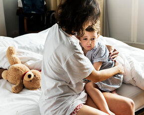 Eine Person umarmt ein Kind auf einem Bett, während ein Teddybär daneben liegt. Das Kind wirkt nachdenklich.