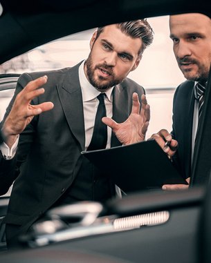 Zwei Männer in Anzügen diskutieren im Innenraum eines Autos. Einer von ihnen gestikuliert, während er ein Tablet oder Notizblock hält. Der andere hört aufmerksam zu und macht sich Notizen.