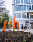 Ein modernes Gebäude des SRH Campus Berlin mit einer auffälligen orangefarbenen Schriftzug "srh" im Vordergrund. Einige Personen stehen vor dem Eingang und unterhalten sich.
