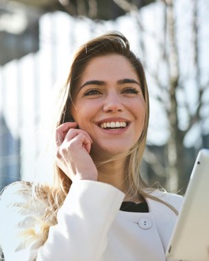 Eine lächelnde Frau mit langen Haaren hält ein Tablet in der Hand und spricht telefonierend. Sie trägt einen weißen Mantel und steht in einer urbanen Umgebung mit modernen Gebäuden im Hintergrund.