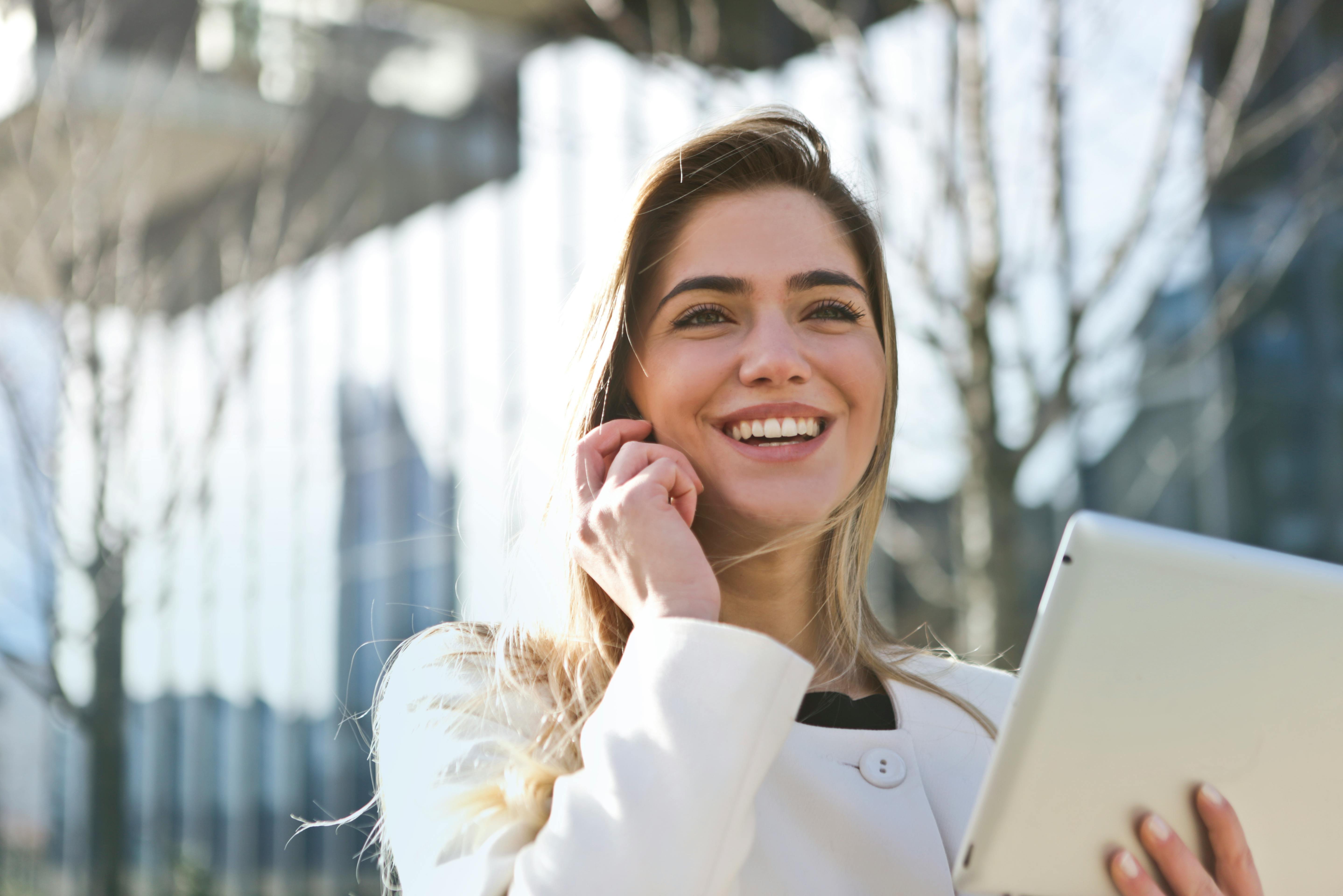 Eine lächelnde Frau mit langen Haaren hält ein Tablet in der Hand und spricht telefonierend. Sie trägt einen weißen Mantel und steht in einer urbanen Umgebung mit modernen Gebäuden im Hintergrund.