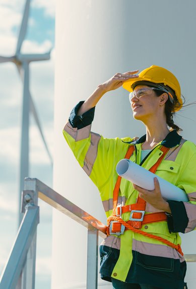 A woman in safety gear and a helmet stands by a wind turbine, looking into the distance while holding plans in her hand.