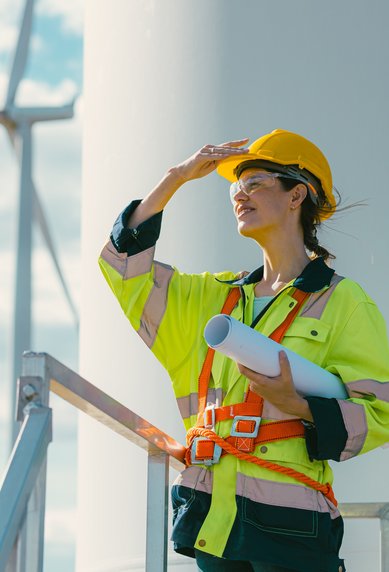 A woman in safety gear and a helmet stands by a wind turbine, looking into the distance while holding plans in her hand.