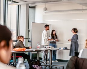 Three people are standing in a classroom discussing. One person is taking notes while another is speaking. In the background, students can be seen at desks with water bottles and materials.