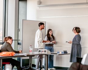 Three people are standing in a classroom discussing. One person is taking notes while another is speaking. In the background, students can be seen at desks with water bottles and materials.