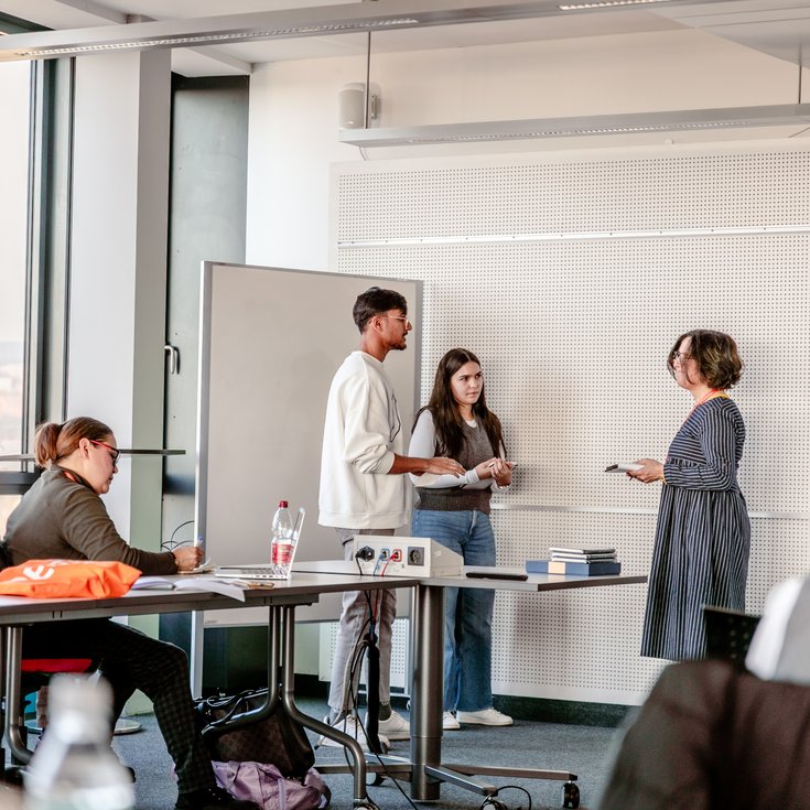 Three people are standing in a classroom discussing. One person is taking notes while another is speaking. In the background, students can be seen at desks with water bottles and materials.