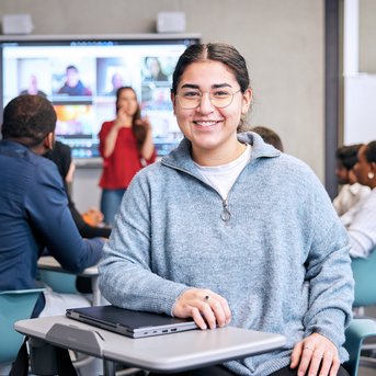 A smiling student sits at a table in a modern classroom, while a presentation is displayed on a screen in the background. Other students are also present and listening attentively.