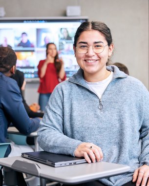 A smiling student sits at a table in a modern classroom, while a presentation is displayed on a screen in the background. Other students are also present and listening attentively.
