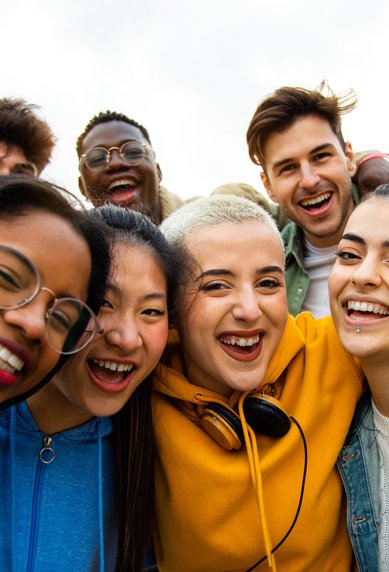 A group of seven young people smiles and happily poses for a selfie outdoors. They are dressed casually and radiate joy.
