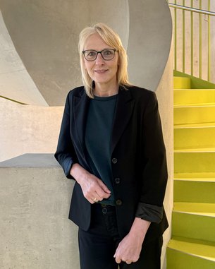 Eine blonde Frau mit Brille steht an einer Betonwand und lächelt, während sie sich an einem Geländer neben einer grünen Treppe lehnt.