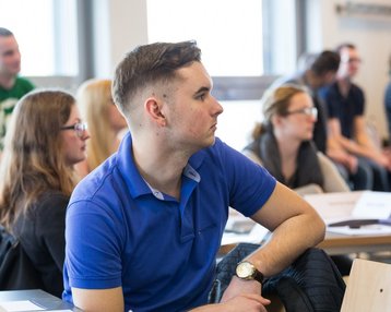 Eine Gruppe von Studierenden hört aufmerksam zu, während ein junger Mann in einem blauen Polohemd nachdenklich schaut.