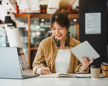 Eine Frau sitzt an einem Tisch, schreibt in ein Notizbuch und hält ein Tablet in der Hand. Ein Laptop und Bücher sind ebenfalls sichtbar.