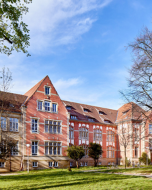 Historic building with a red facade, surrounded by trees and a lawn, under a blue sky.