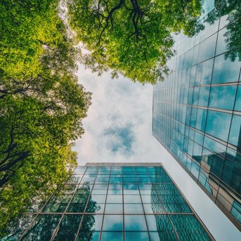 Looking up at modern glass buildings, surrounded by tall trees and a partly cloudy sky.