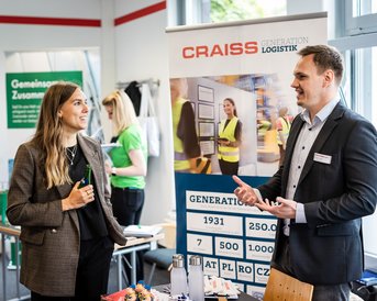 Zwei Personen im Gespräch an einem Messestand, mit einem Banner der Firma CRAISS im Hintergrund, das Informationen zur Logistik bietet.