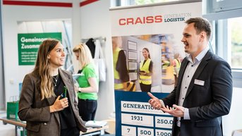 Zwei Personen im Gespräch an einem Messestand, mit einem Banner der Firma CRAISS im Hintergrund, das Informationen zur Logistik bietet.