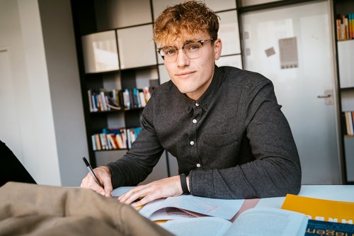 Ein junger Mann mit lockigem Haar und Brille sitzt an einem Tisch, schreibt in ein Notizbuch und hat Bücher vor sich.