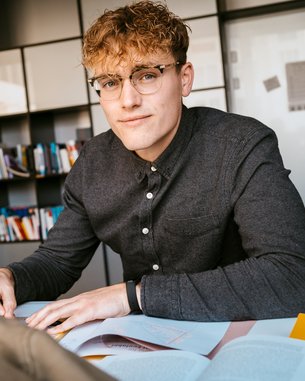 Ein junger Mann mit lockigem Haar und Brille sitzt an einem Tisch, schreibt in ein Notizbuch und hat Bücher vor sich.