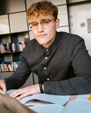 A young man with curly hair and glasses is sitting at a table, writing in a notebook, with books in front of him.