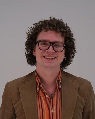 Ein lächelnder Mann mit lockigem Haar trägt eine Brille und ein braunes Sakko über einem gestreiften Hemd.