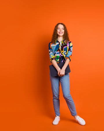 Eine junge Frau mit lockigem Haar trägt ein buntes, gemustertes Hemd und Jeans. Sie steht vor einem orangefarbenen Hintergrund und hält einen Laptop in der Hand. Ihr Gesicht zeigt ein freundliches Lächeln.