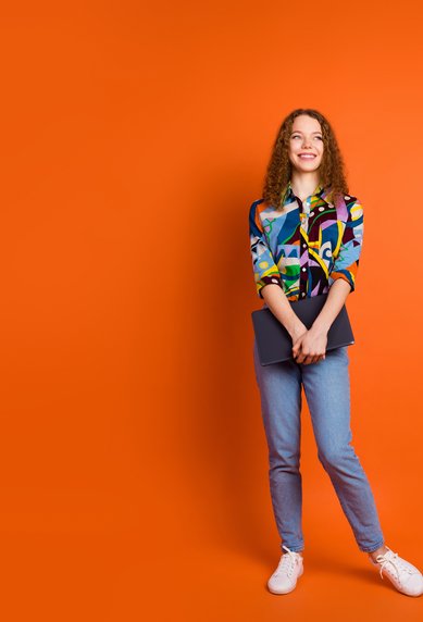 A young woman with curly hair is wearing a colorful, patterned shirt and jeans. She stands in front of an orange background, holding a laptop in her hand. Her face displays a friendly smile.