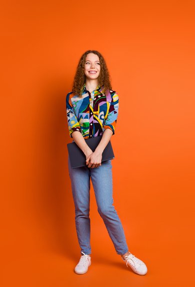 A young woman with curly hair is wearing a colorful, patterned shirt and jeans. She stands in front of an orange background, holding a laptop in her hand. Her face displays a friendly smile.