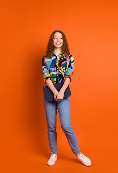 Eine junge Frau mit lockigem Haar trägt ein buntes, gemustertes Hemd und Jeans. Sie steht vor einem orangefarbenen Hintergrund und hält einen Laptop in der Hand. Ihr Gesicht zeigt ein freundliches Lächeln.