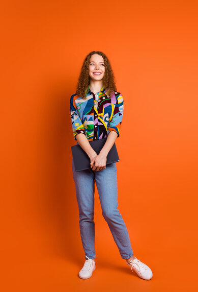 A young woman with curly hair is wearing a colorful, patterned shirt and jeans. She stands in front of an orange background, holding a laptop in her hand. Her face displays a friendly smile.