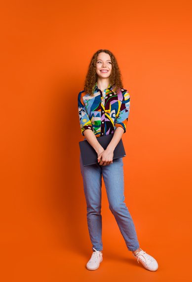 A young woman with curly hair is wearing a colorful, patterned shirt and jeans. She stands in front of an orange background, holding a laptop in her hand. Her face displays a friendly smile.