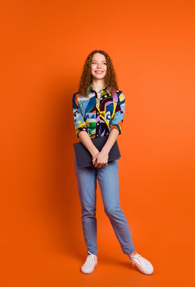 Eine junge Frau mit lockigem Haar trägt ein buntes, gemustertes Hemd und Jeans. Sie steht vor einem orangefarbenen Hintergrund und hält einen Laptop in der Hand. Ihr Gesicht zeigt ein freundliches Lächeln.