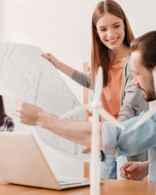 Zwei Personen besprechen Baupläne am Tisch, während eine dritte Person im Hintergrund am Laptop arbeitet. Auf dem Tisch stehen Miniatur-Windkraftanlagen. Die Atmosphäre ist kreativ und kollaborativ.