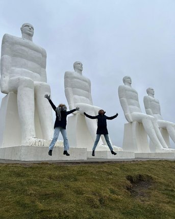 Zwei Personen springen vor vier großen, weißen Skulpturen sitzender Figuren. Der Himmel ist bewölkt und die Umgebung ist grün.