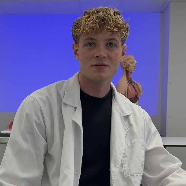 Ein junger Mann in einem weißen Laborkittel sitzt in einem Klassenzimmer mit einem anatomischen Modell im Hintergrund.