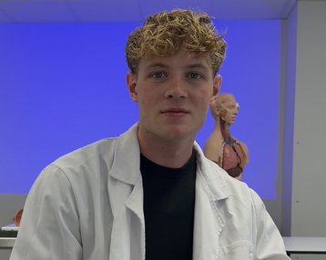 Ein junger Mann in einem weißen Laborkittel sitzt in einem Klassenzimmer mit einem anatomischen Modell im Hintergrund.