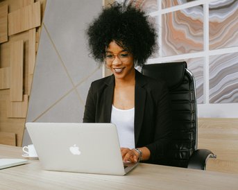 Eine Frau mit lockigem Haar sitzt an einem Schreibtisch und arbeitet an einem Laptop. Sie trägt eine schwarze Blazerjacke und eine Brille, während sie lächelt und auf den Bildschirm schaut.