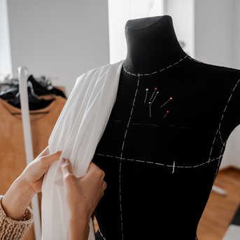A person drapes a white fabric over a dress form that is pinned with sewing pins. In the background, clothing items are visible.