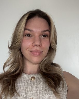 Eine Person mit langen, welligen Haaren und einem beigen Oberteil steht vor einer weißen Wand und lächelt leicht.