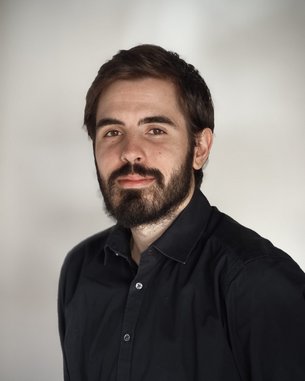 A man with a beard is wearing a black shirt and looking directly at the camera. The background is neutral and bright.