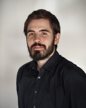 Ein Mann mit Bart und dunklem Hemd steht vor einem hellen Hintergrund und schaut direkt in die Kamera.