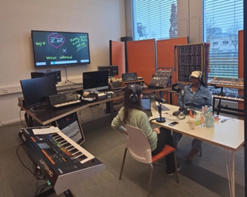 Two people are sitting at a table in a modern studio, surrounded by musical instruments and computers, while they engage in a conversation.