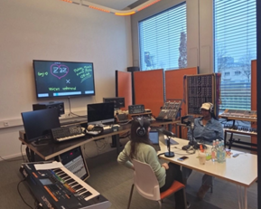 Two people are sitting at a table in a modern studio, surrounded by musical instruments and computers, while they engage in a conversation.