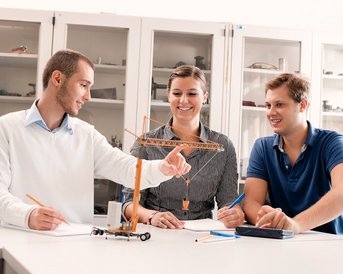 Drei Personen sitzen an einem Tisch und diskutieren. Eine Person zeigt auf ein Modell eines Krans, während die anderen aufmerksam zuhören und Notizen machen.