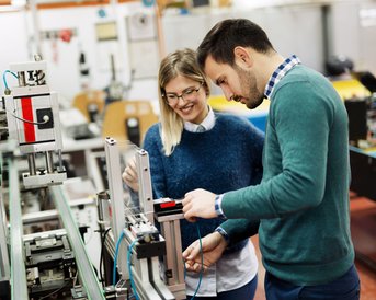 Zwei Personen in einem modernen Labor arbeiten zusammen an einer Maschine. Die Frau trägt eine Brille und lächelt, während der Mann konzentriert eine Komponente überprüft.