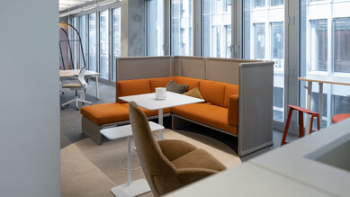 A modern office with large windows, a cozy orange sofa, a table, and a chair. Bright colors and an open layout create an inviting atmosphere.