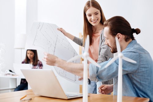 Zwei Personen diskutieren Pläne vor einem Laptop, während eine dritte Person im Hintergrund am Computer arbeitet. Windkraftanlagen sind sichtbar.