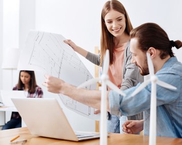 Zwei Personen diskutieren Pläne vor einem Laptop, während eine dritte Person im Hintergrund am Computer arbeitet. Windkraftanlagen sind sichtbar.